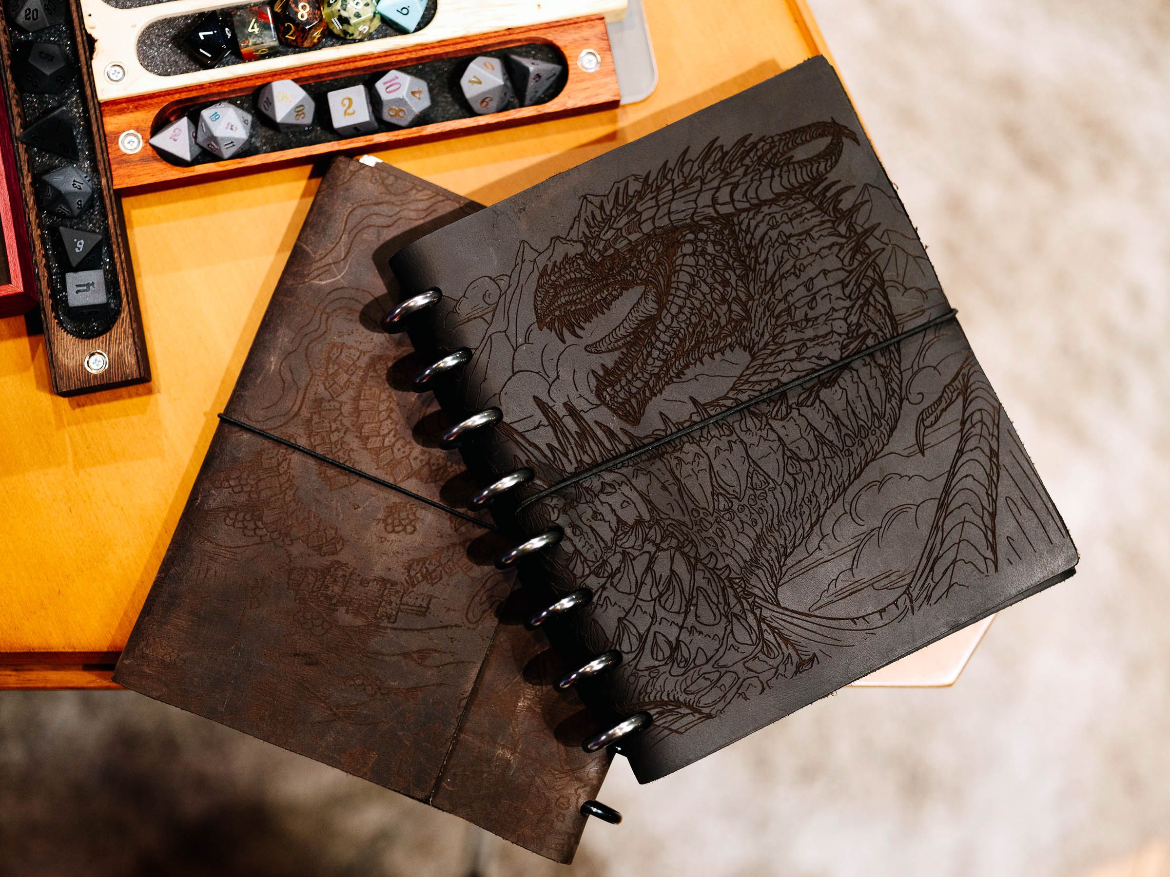 A photo of a leather-bound notebook with a dragon etched on the cover. Above it are other tools for playing Dungeons and Dragons, including a few sets of dice.